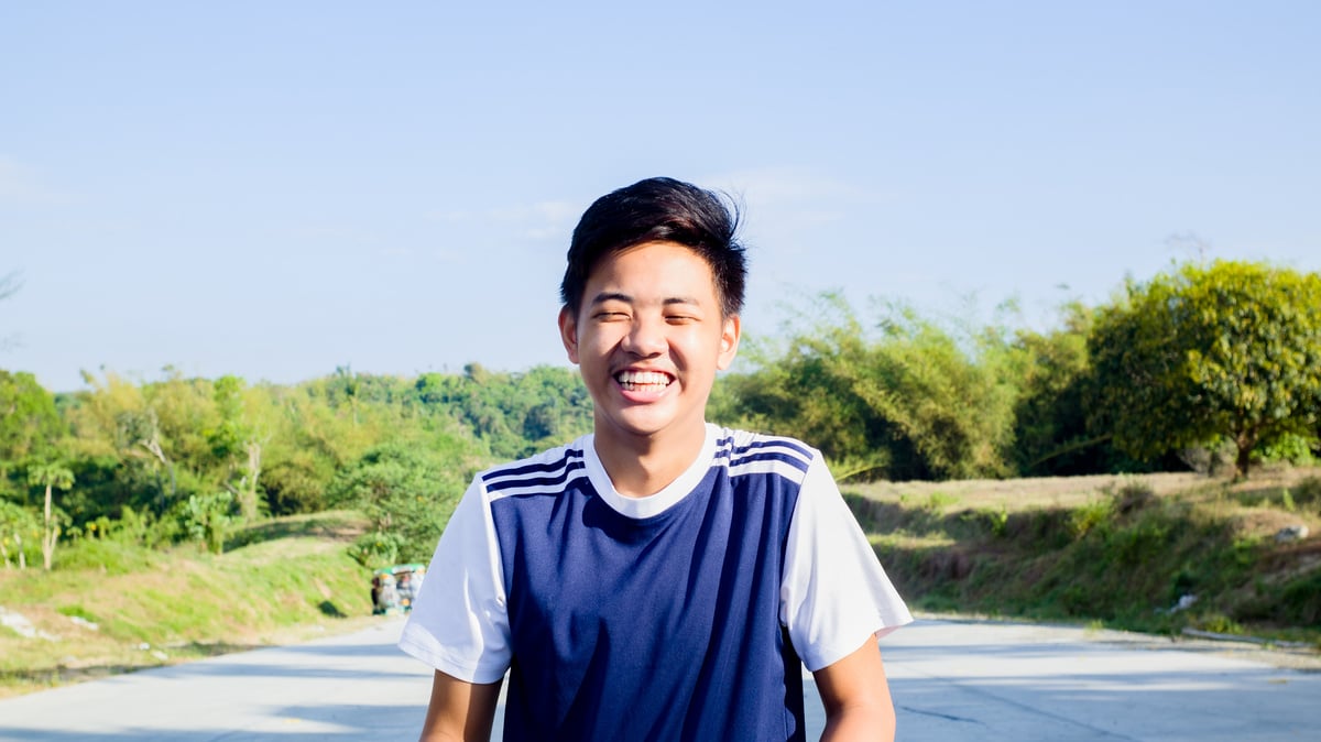 Happy Asian guy standing on roadway against green trees in sunlight